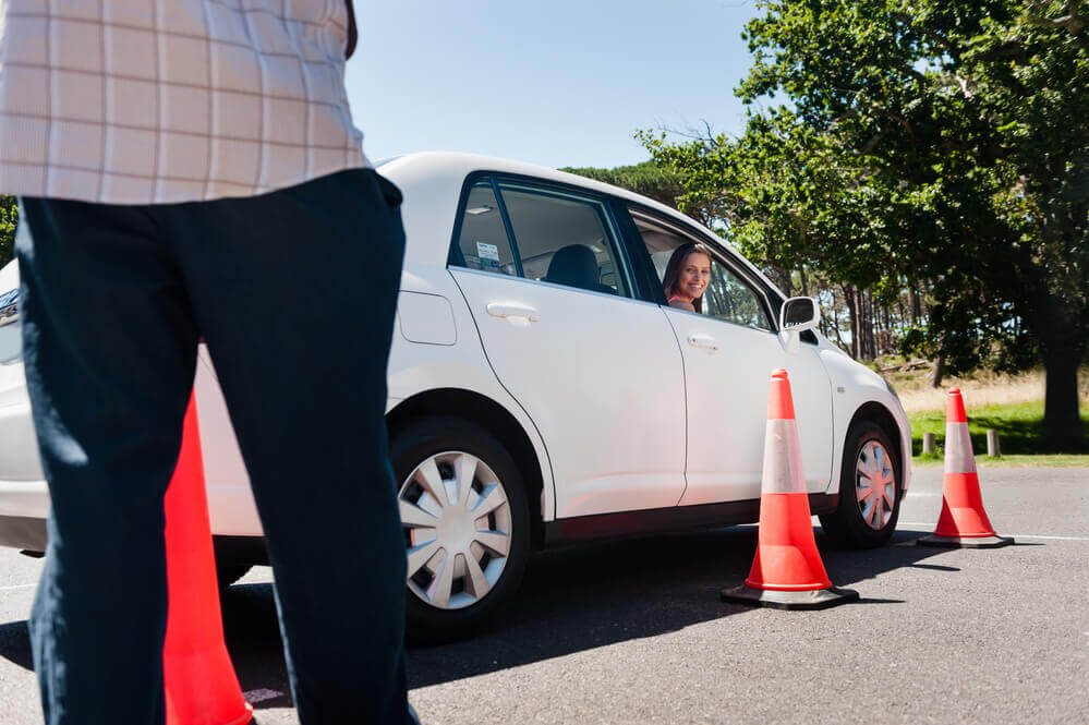 A driving school Mandurah instructor assessing a learner park inside of some red cones while he stands outside the car with a clip board